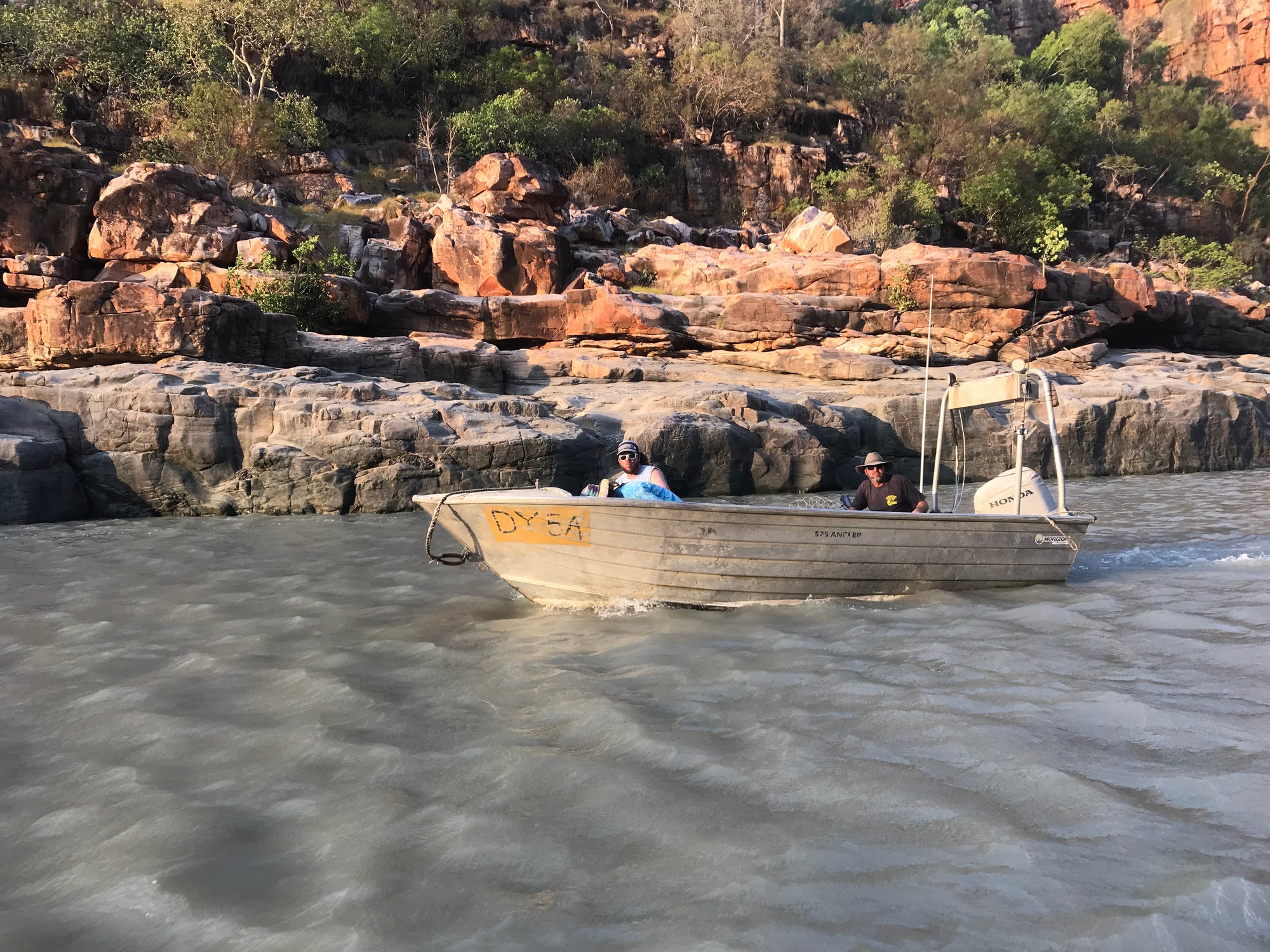 Two people in a boat on a river with rocky cliffs and trees in the background