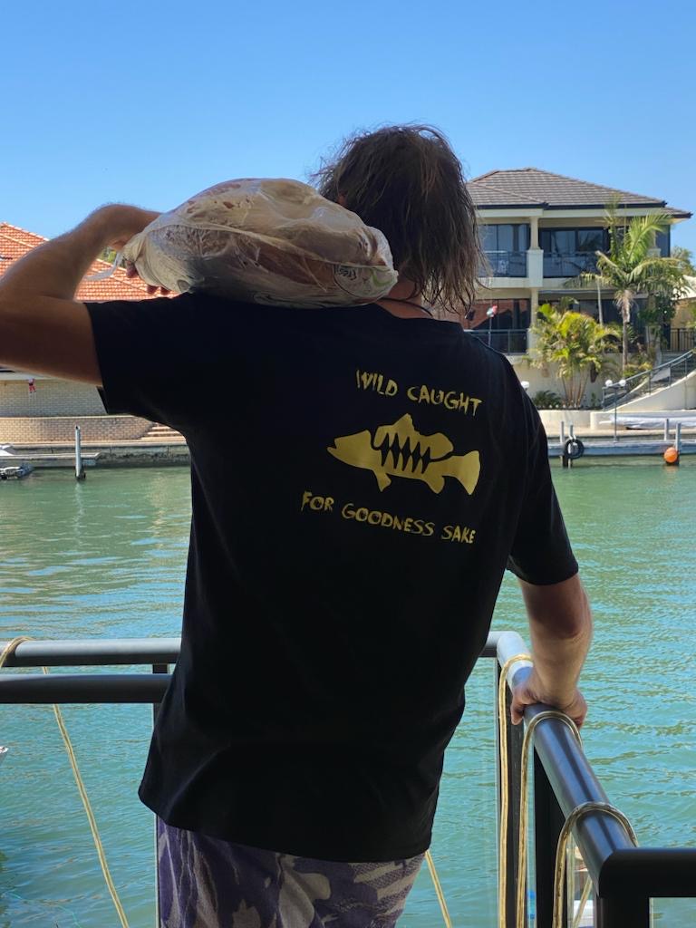 Person wearing a black t-shirt with a fish graphic and text by a waterfront.
